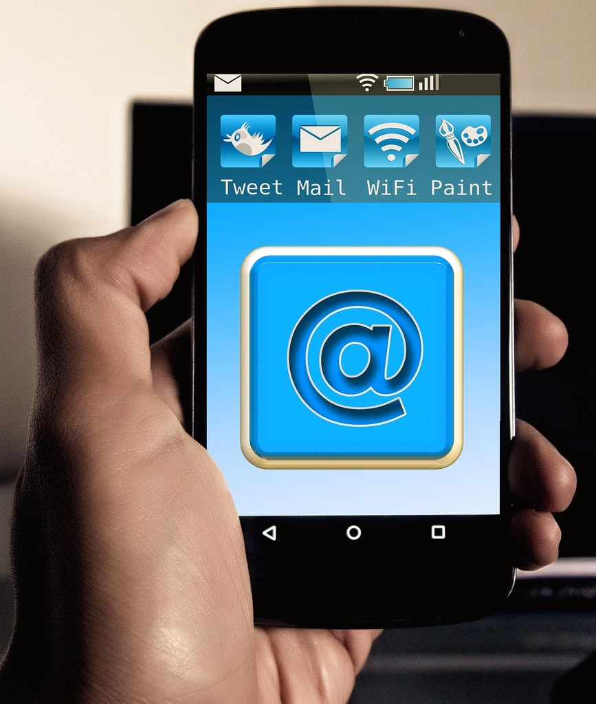 Gros plan d'une main tenant un smartphone affichant une application avec l'icône '@', symbolisant l'accès à l'email et autres fonctions connectées